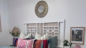Un dormitorio ordenado con cama, una mesita de noche con un florero y un cuadro enmarcado, y un espejo decorativo en la pared.
