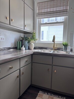 Een keuken met grijze kastjes, een marmeren aanrecht, een spoelbak en een raam met zonwering.