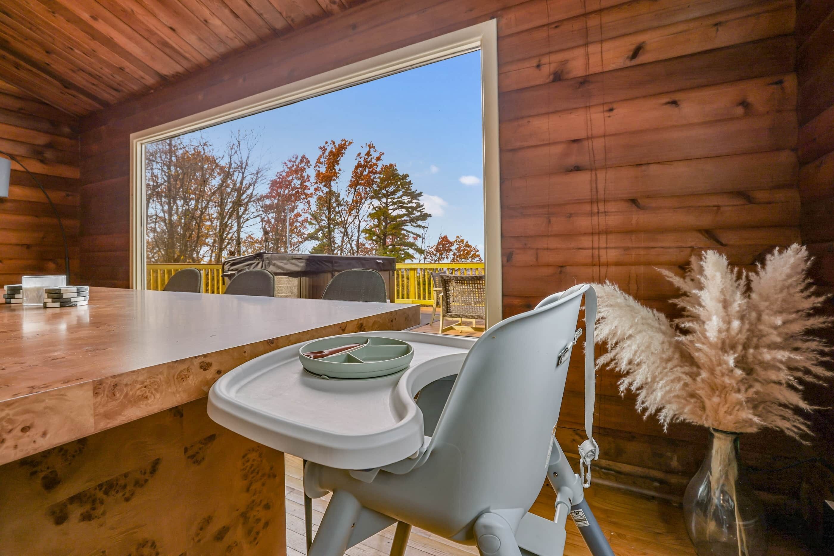 A wooden cabin interior with a dining table, a high chair, and a view of trees through a large window.