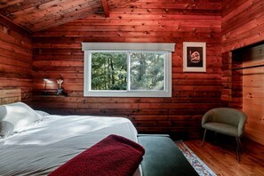 Une pièce de style cabane en bois, avec un lit, une chaise, une fenêtre donnant sur les arbres et un tableau encadré au mur.