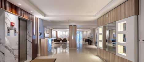 A hotel corridor with marble walls, wooden paneling, a reception desk, seating area, and a view into the room.