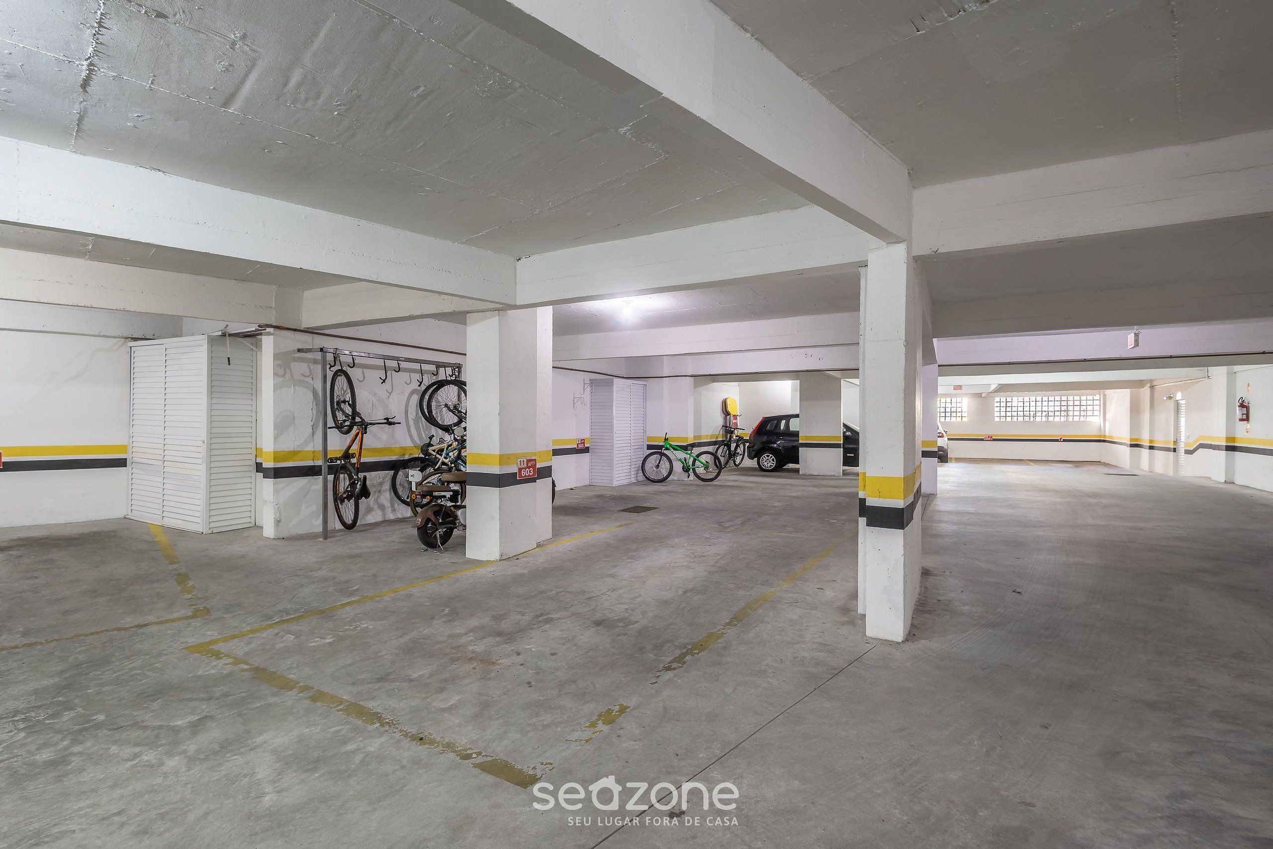 Un estacionamiento vacío con pisos de concreto y paredes blancas, con líneas a rayas amarillas y negras, y varias bicicletas estacionadas contra las paredes.