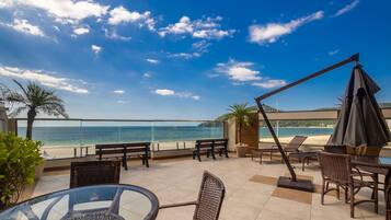 A terrace with a view of the sea, furnished with wicker chairs and tables, and a glass balustrade.