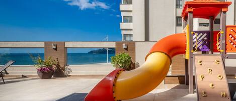 Zona de juegos infantiles con tobogán, pared de escalar y vista al mar.