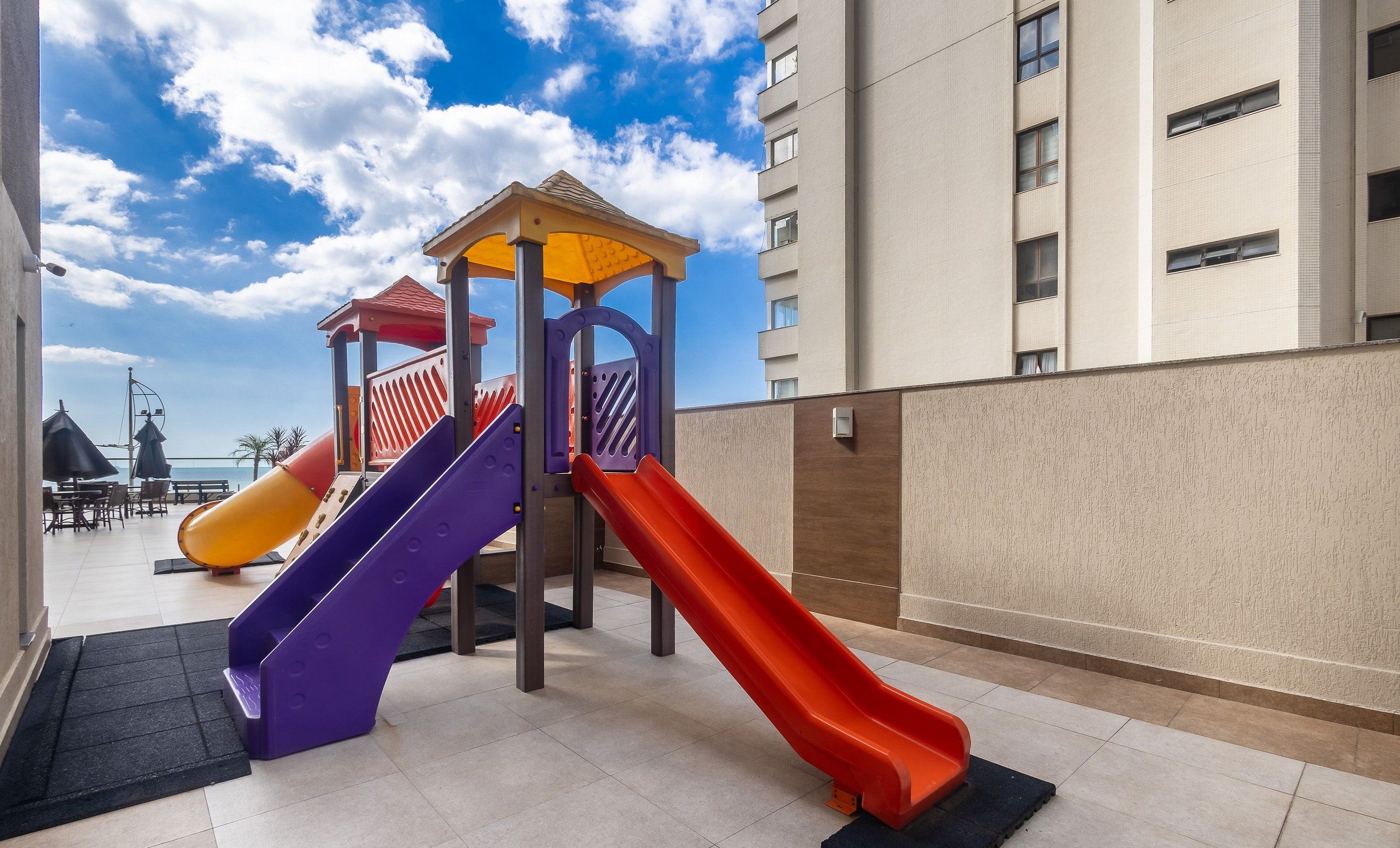 Zona de juegos infantiles con toboganes y un pequeño refugio, ubicada en un edificio residencial con vistas al mar.