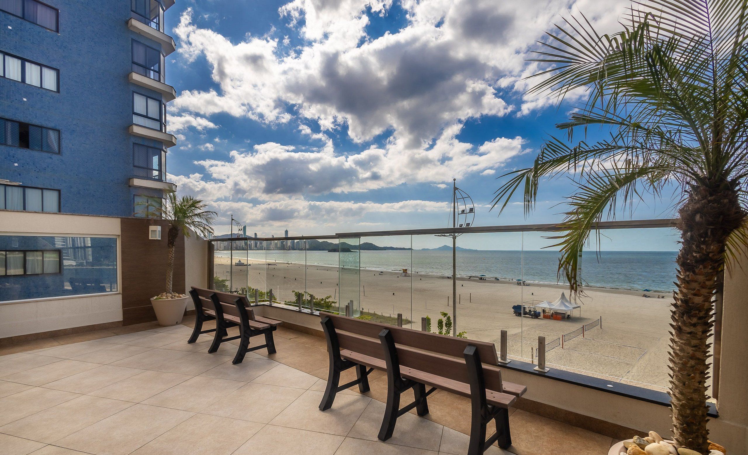 A balcony with a view of a beach, a palm tree, and a building in the background.