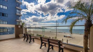A balcony with a view of a beach, a palm tree, and a building in the background.