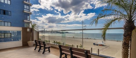 A balcony with a view of a beach, a palm tree, and a building in the background.