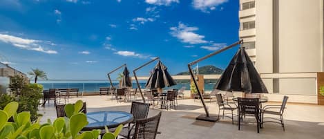 An outdoor seating area with tables and chairs, umbrellas, and a view of the sea.