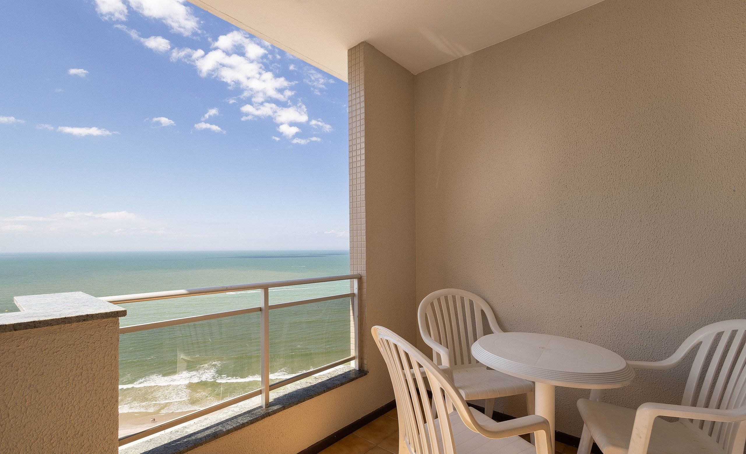 A balcony with a view of the ocean, a small round table with chairs, and a tiled wall.