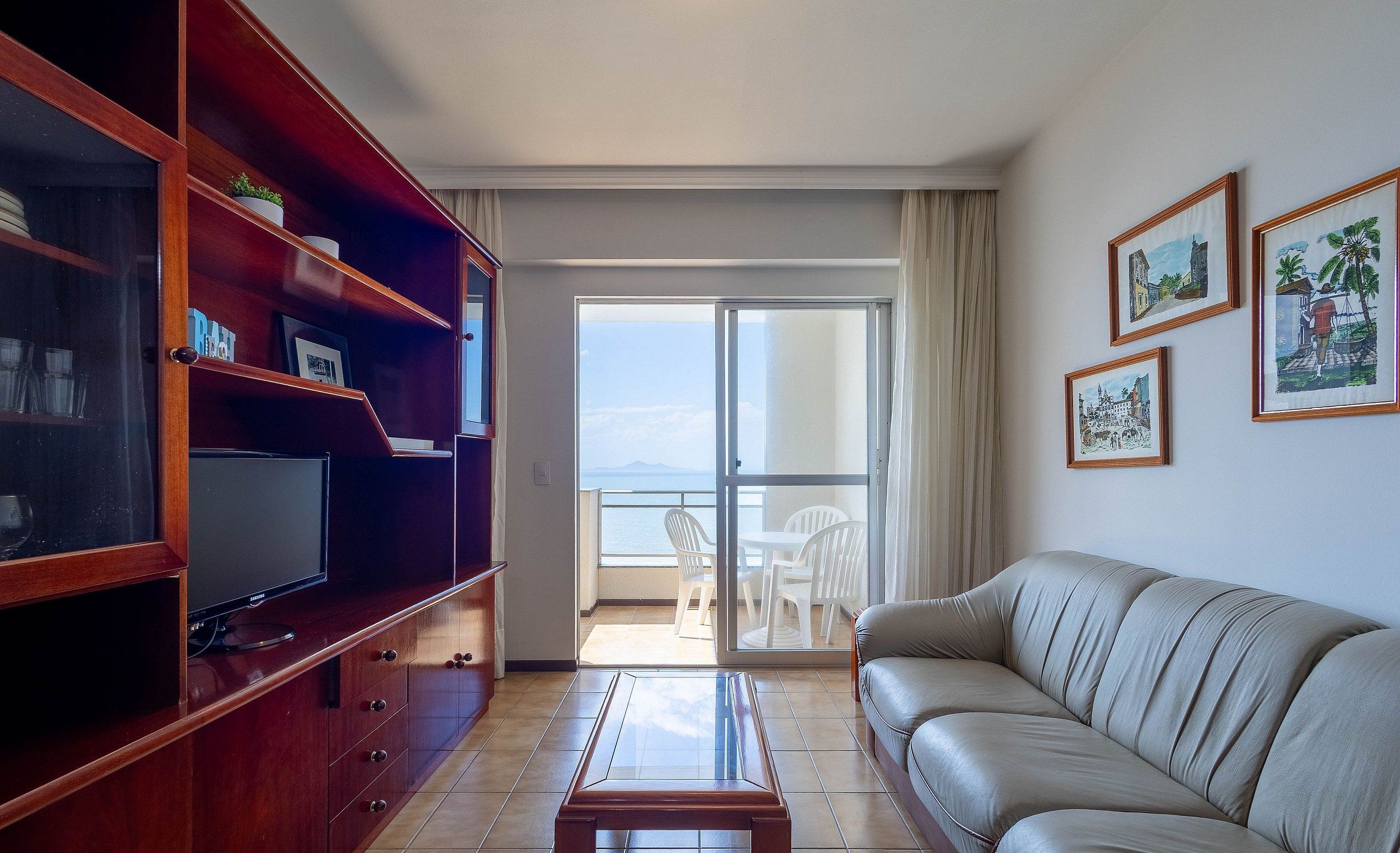 Uma sala de estar com sofá, televisão em um aparador de madeira, mesa de centro de vidro e varanda com vista.