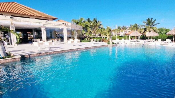 A swimming pool with lounge chairs and umbrellas, surrounded by palm trees and a building with a tiled roof.