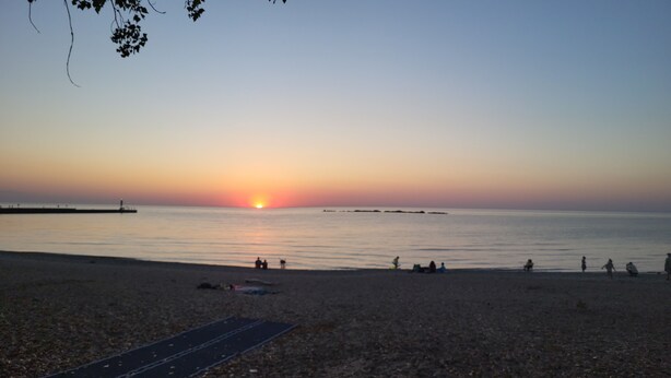 Une plage au coucher du soleil, avec une eau calme, une jetée au loin et des gens qui se détendent sur le sable.