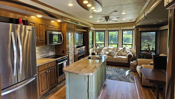A spacious kitchen with a central island, wooden cabinets, stainless steel appliances, and large windows offering views of greenery. - Camper on peaceful gated property. (North Fort Myers)