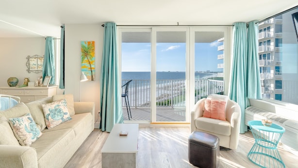A living room with a sofa, coffee table, and armchair, a balcony with a view of the sea, and a large window with curtains.