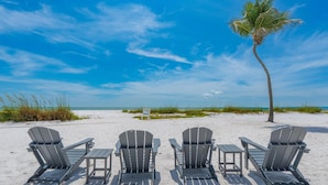 A beach with four Adirondack chairs and a table, a palm tree, and a clear blue sky.