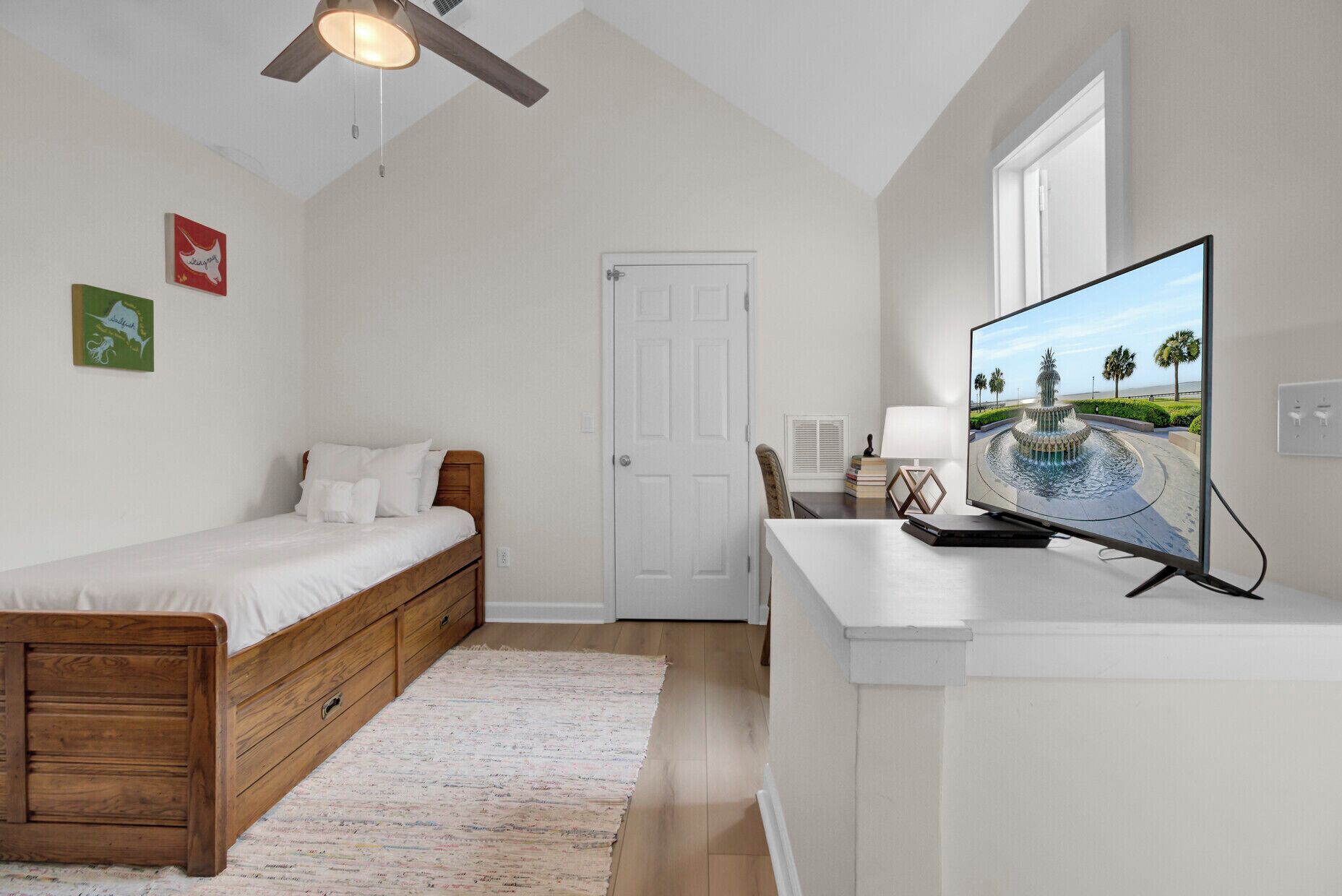 A bedroom with a bed, a desk with a TV, a ceiling fan, and a door.