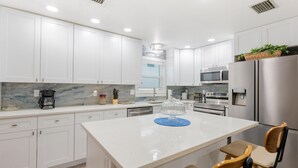 Une cuisine moderne avec des meubles blancs, un îlot central, des appareils électroménagers en acier inoxydable et une fenêtre avec des stores.
