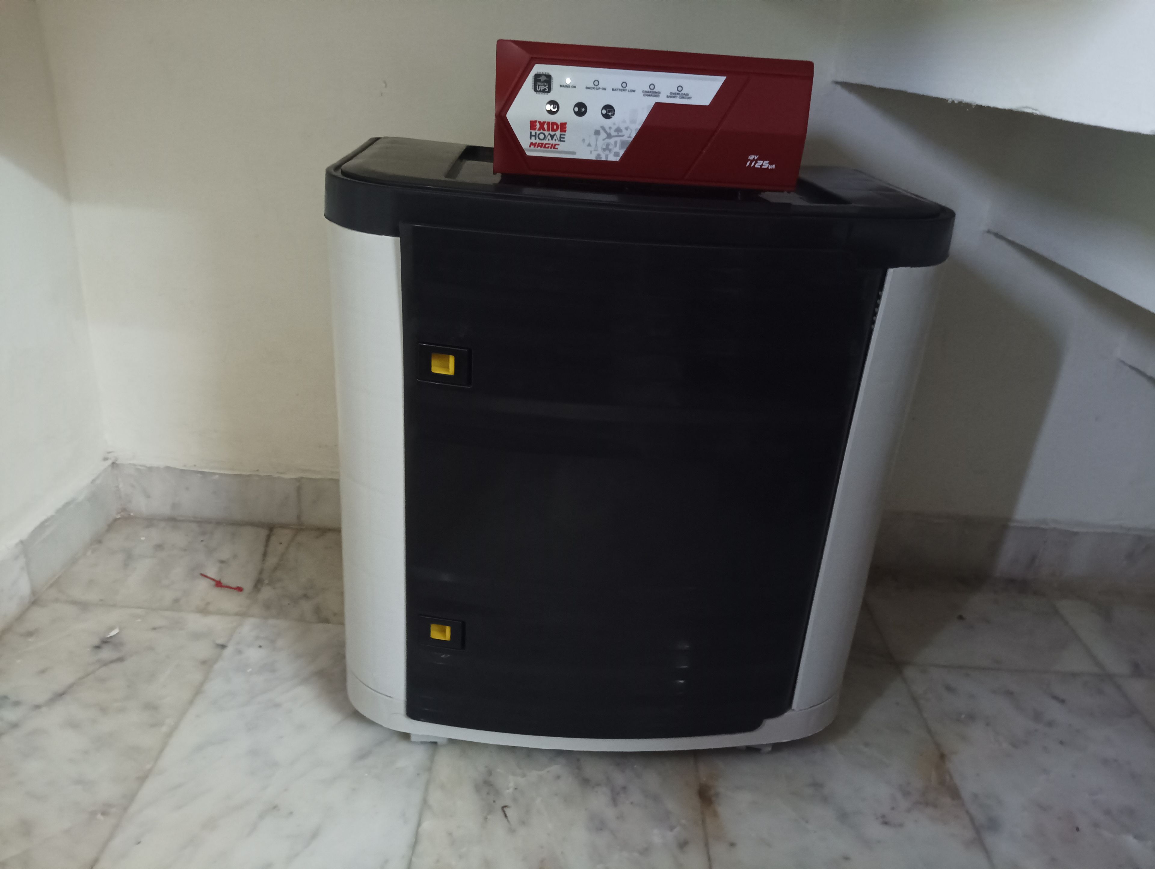 A compact, cylindrical trash bin with a red top and a black front panel, placed on a tiled floor against a wall.