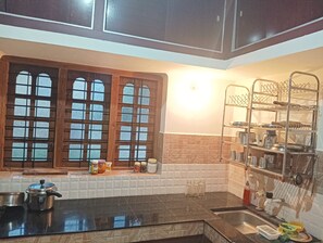 A kitchen with a black countertop, a sink, a window with wooden frames, and a metal rack with kitchenware. - Simple cozy villa retreat within traditionally reputed area on Palakkad, kerala. (Pudunagaram)