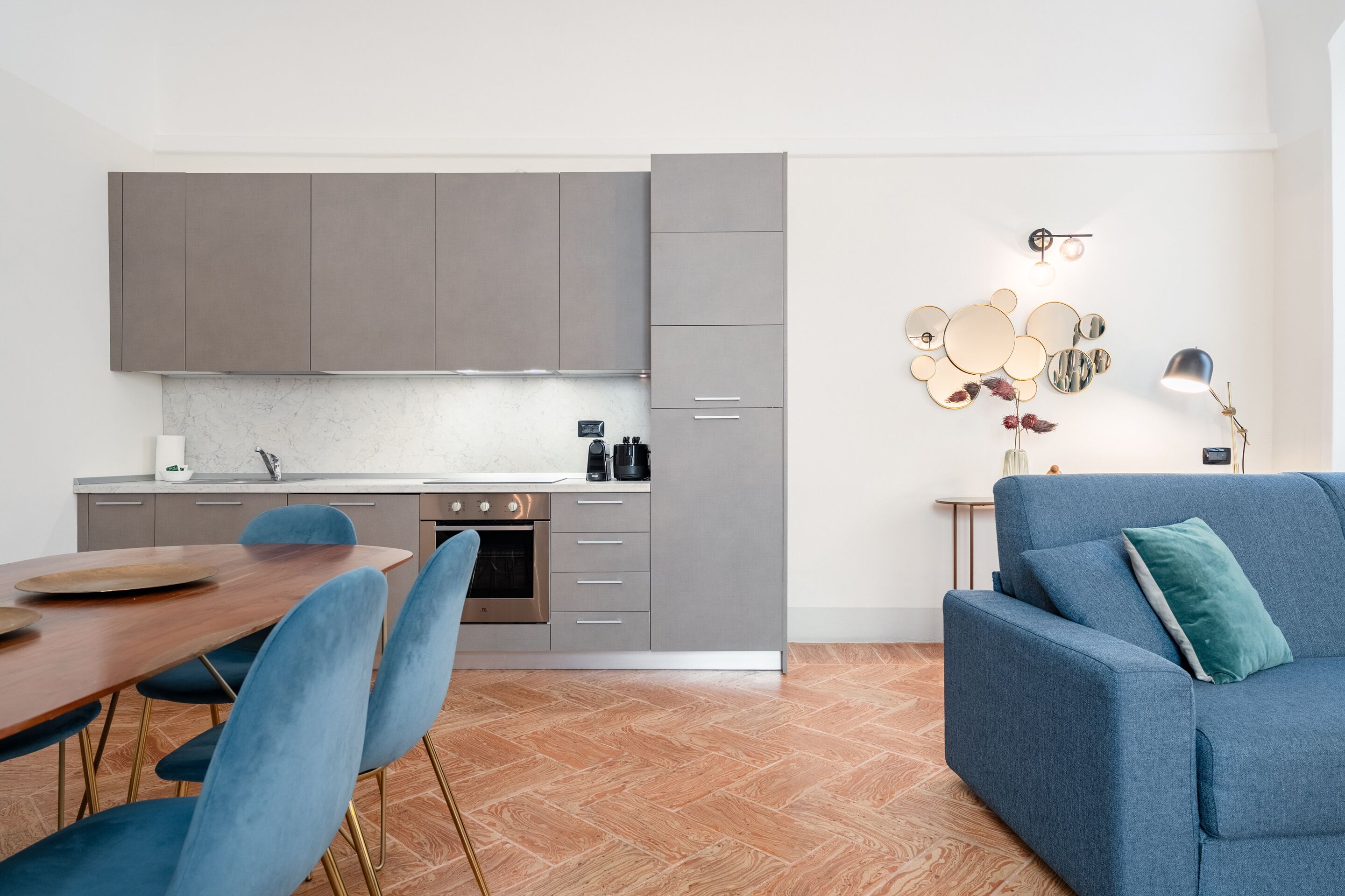 Una cucina moderna con mobili grigi, un tavolo da pranzo in legno con sedie blu e un divano blu in zona living.