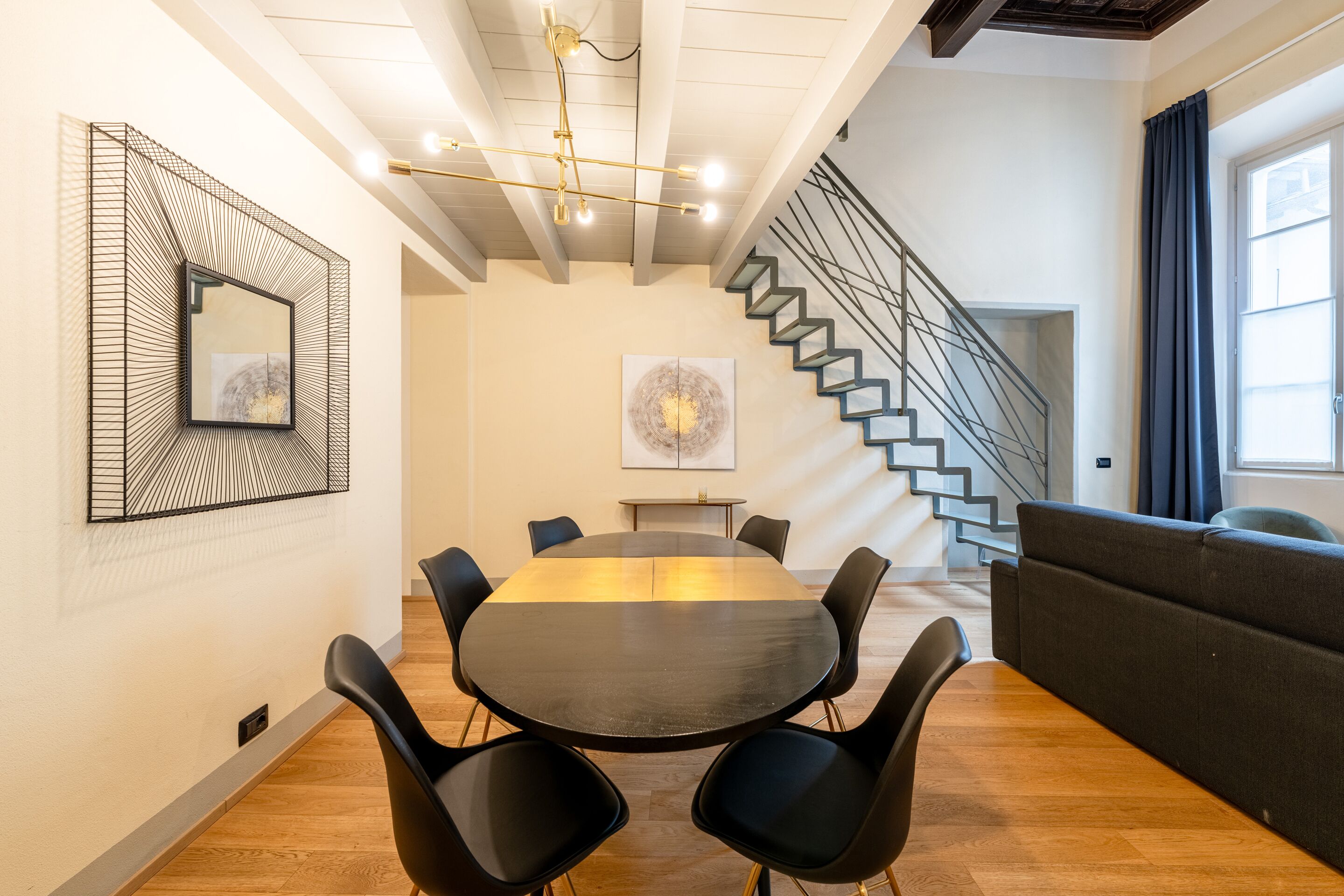 A modern conference room with a round table, black chairs, a large abstract painting, a staircase, and a sofa.