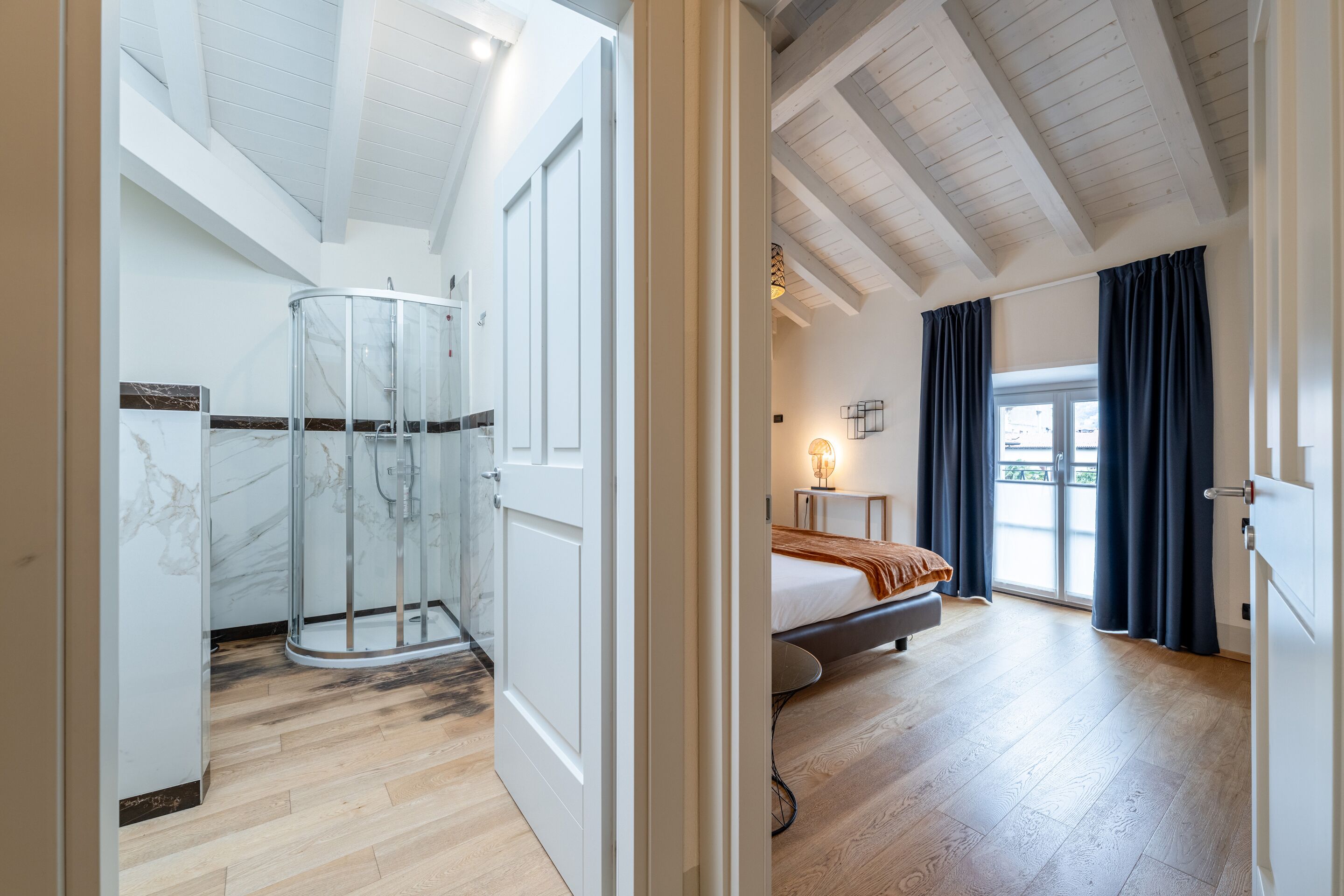 A spacious bedroom with a bed, a bedside table, a window with curtains, and a glass shower enclosure.
