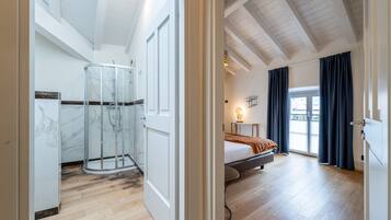 A spacious bedroom with a bed, a bedside table, a window with curtains, and a glass shower enclosure.