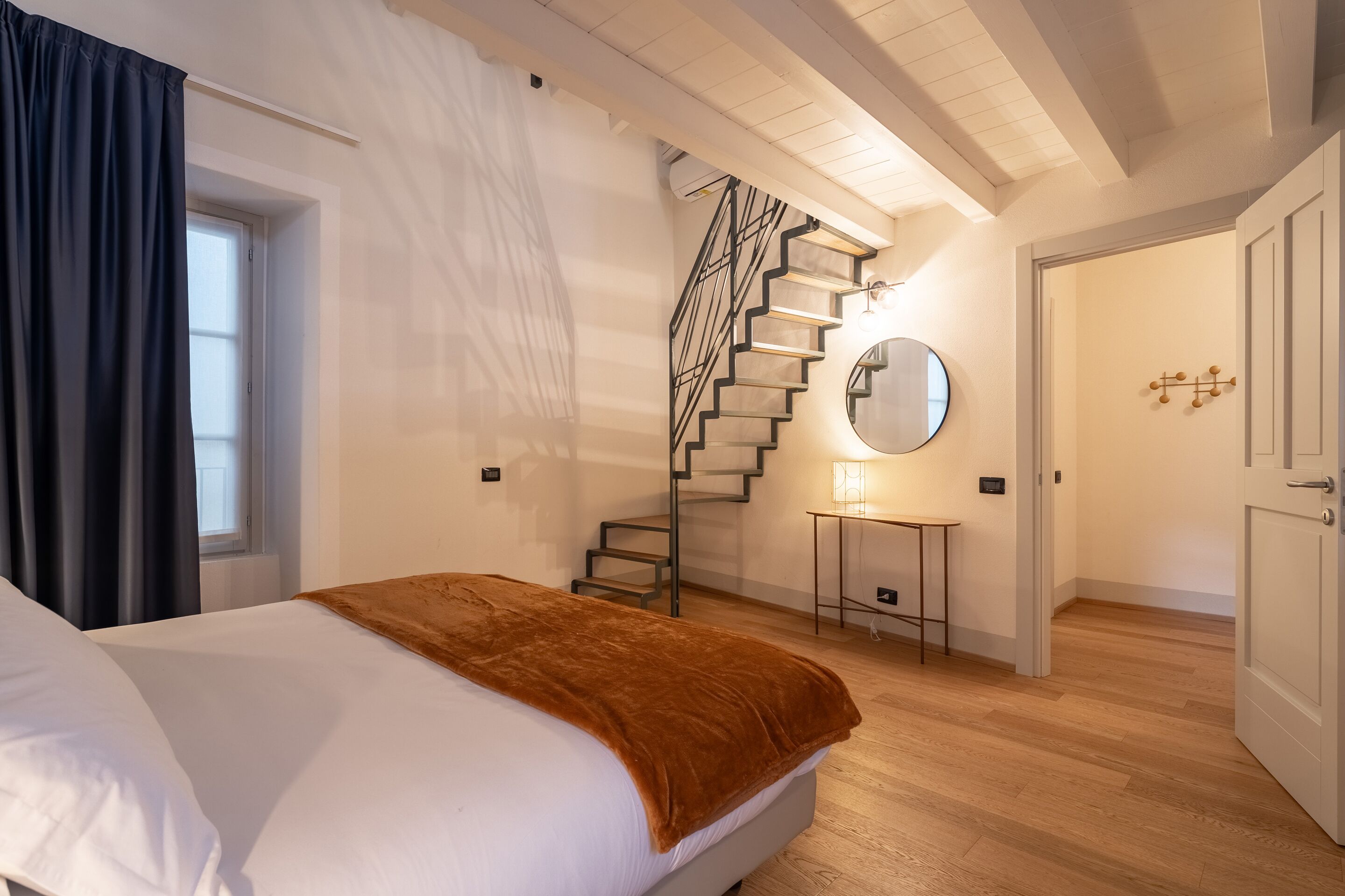 A bedroom with a bed, a staircase, a small table with a lamp, and a door.