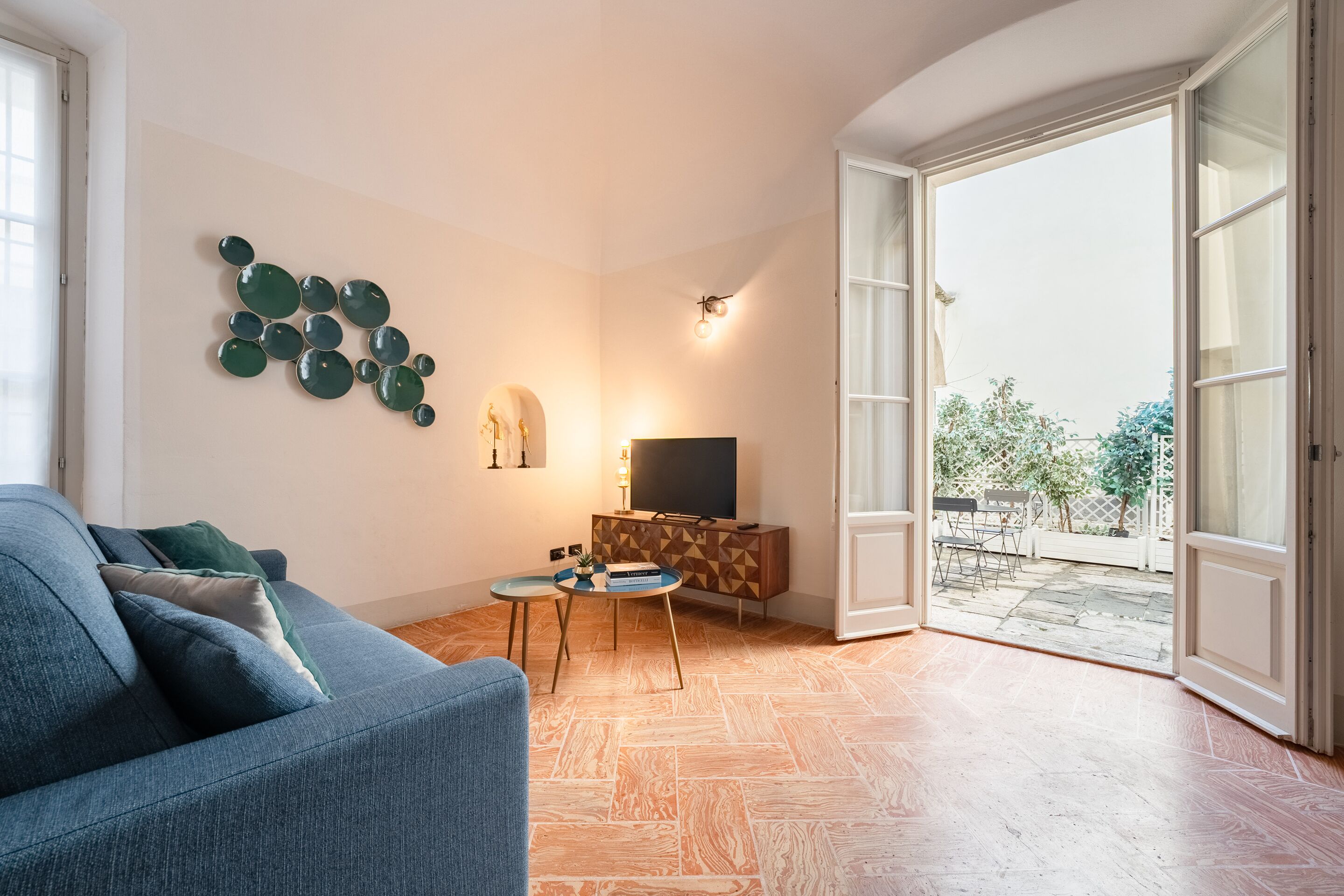 Ein Wohnzimmer mit einer Couch, einem Fernseher auf einer Holzkommode, einem Wandbild und Blick ins Freie durch eine offene Tür.