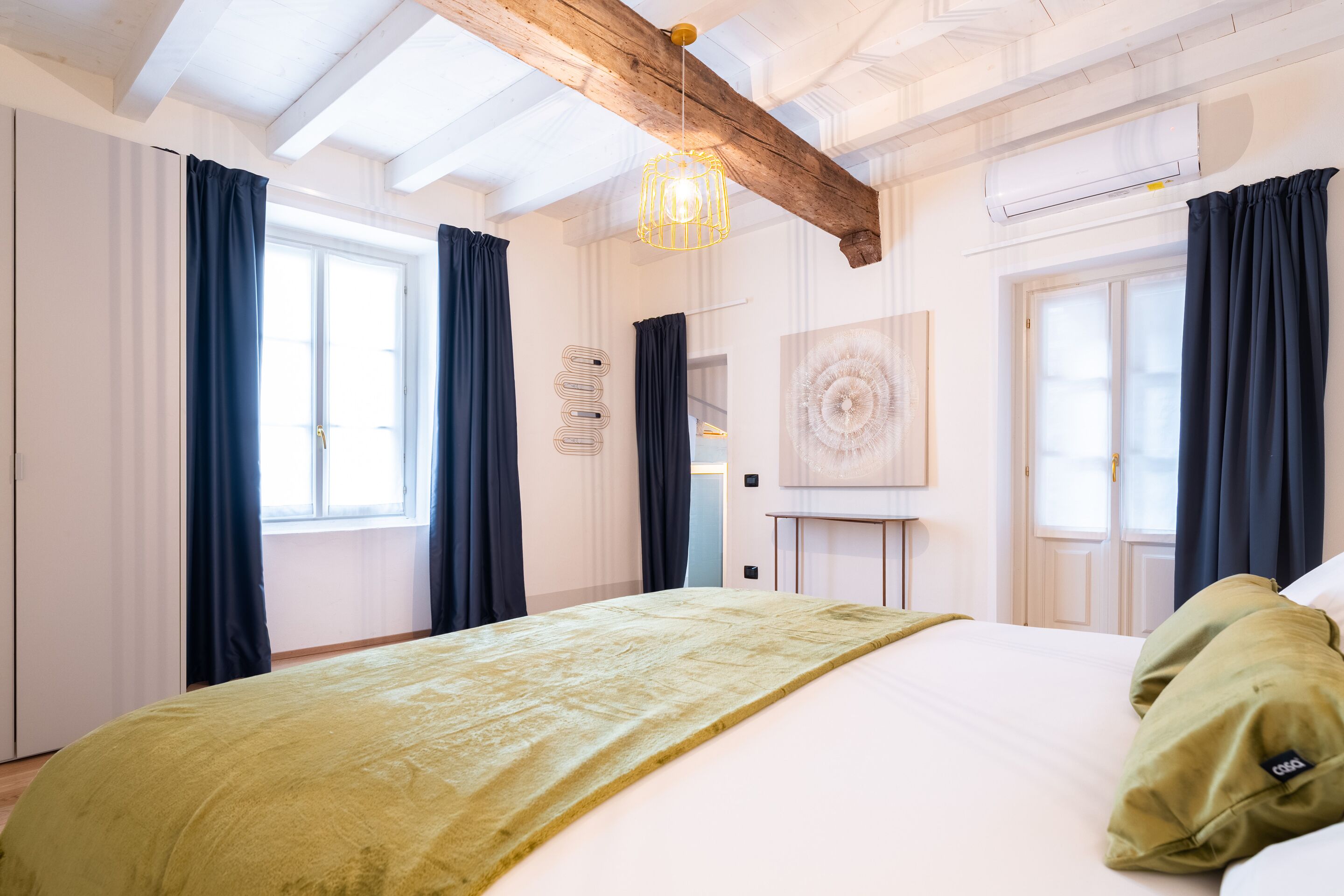 Une chambre d’hôtel avec un grand lit, des rideaux occultants, une poutre en bois au plafond et un luminaire suspendu moderne.