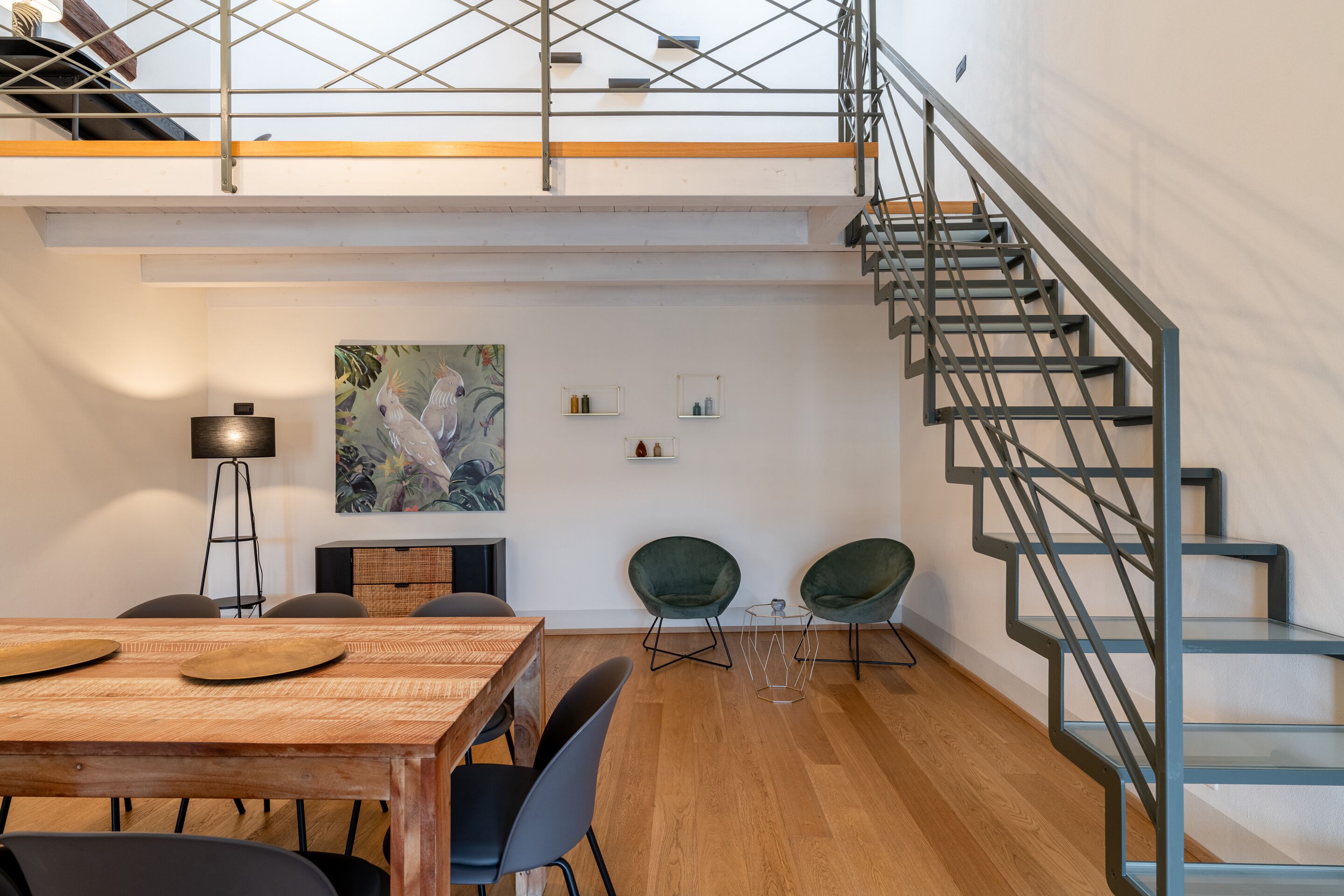 Ein modernes Interieur mit einem Holzes Esstisch, zwei grünen Stühlen, einer Treppe mit Metallgeländern und einem Gemälde an der Wand.