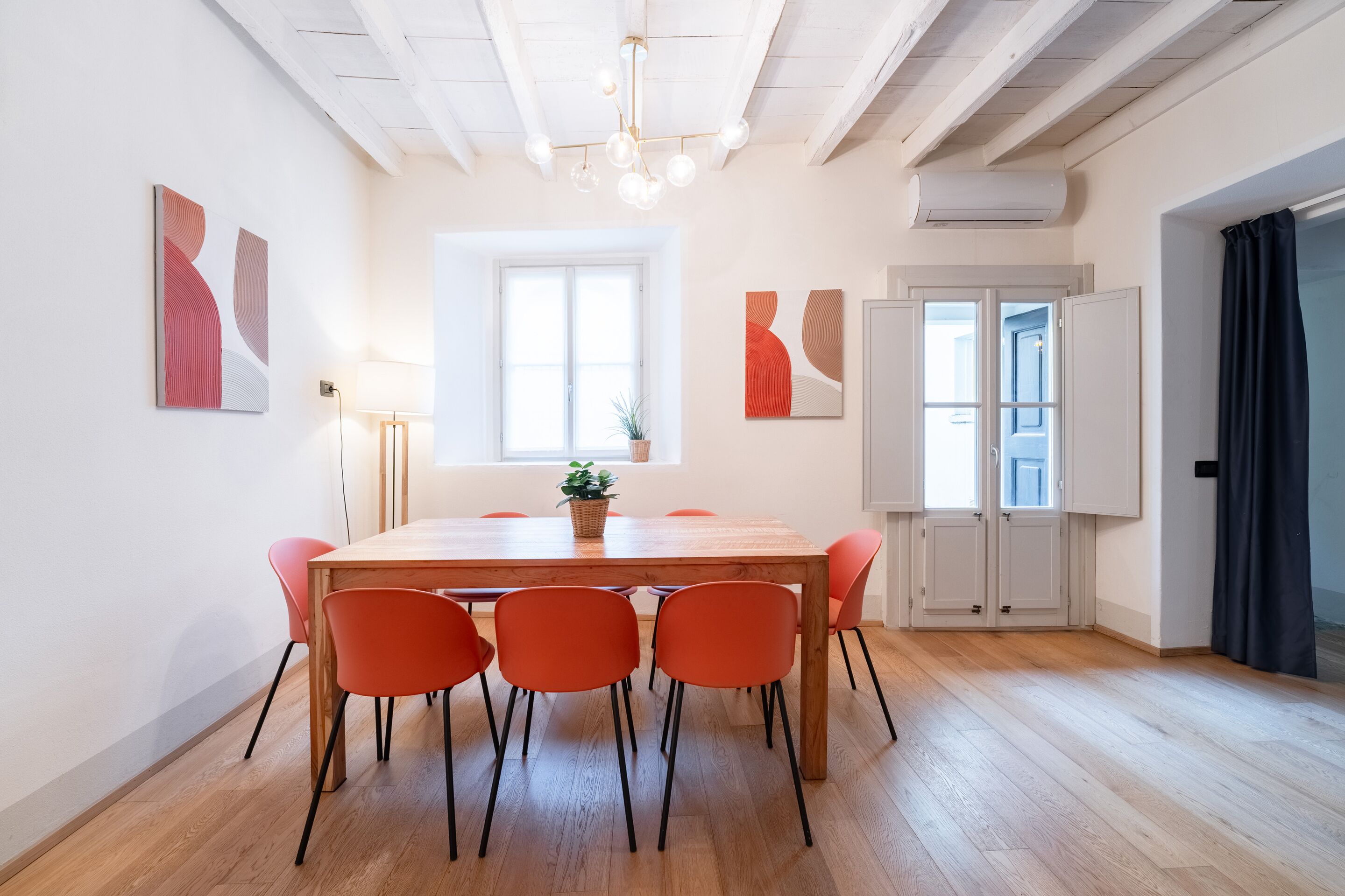 Een eetgedeelte met een houten tafel en oranje stoelen, een plant in pot en abstracte wanddecoratie.