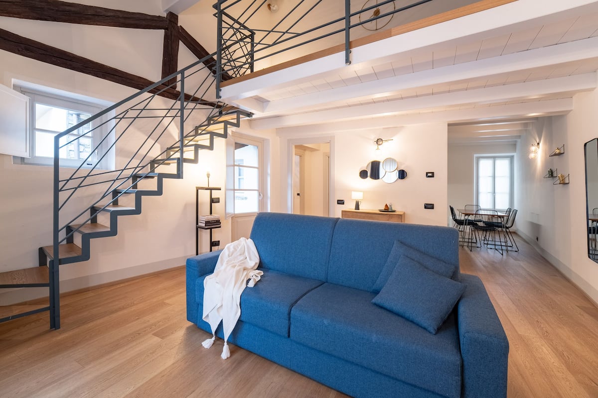Un vaste espace de vie comprenant un canapé bleu, un plancher en bois, un escalier avec des rampes en métal et un coin repas avec une table et des chaises.