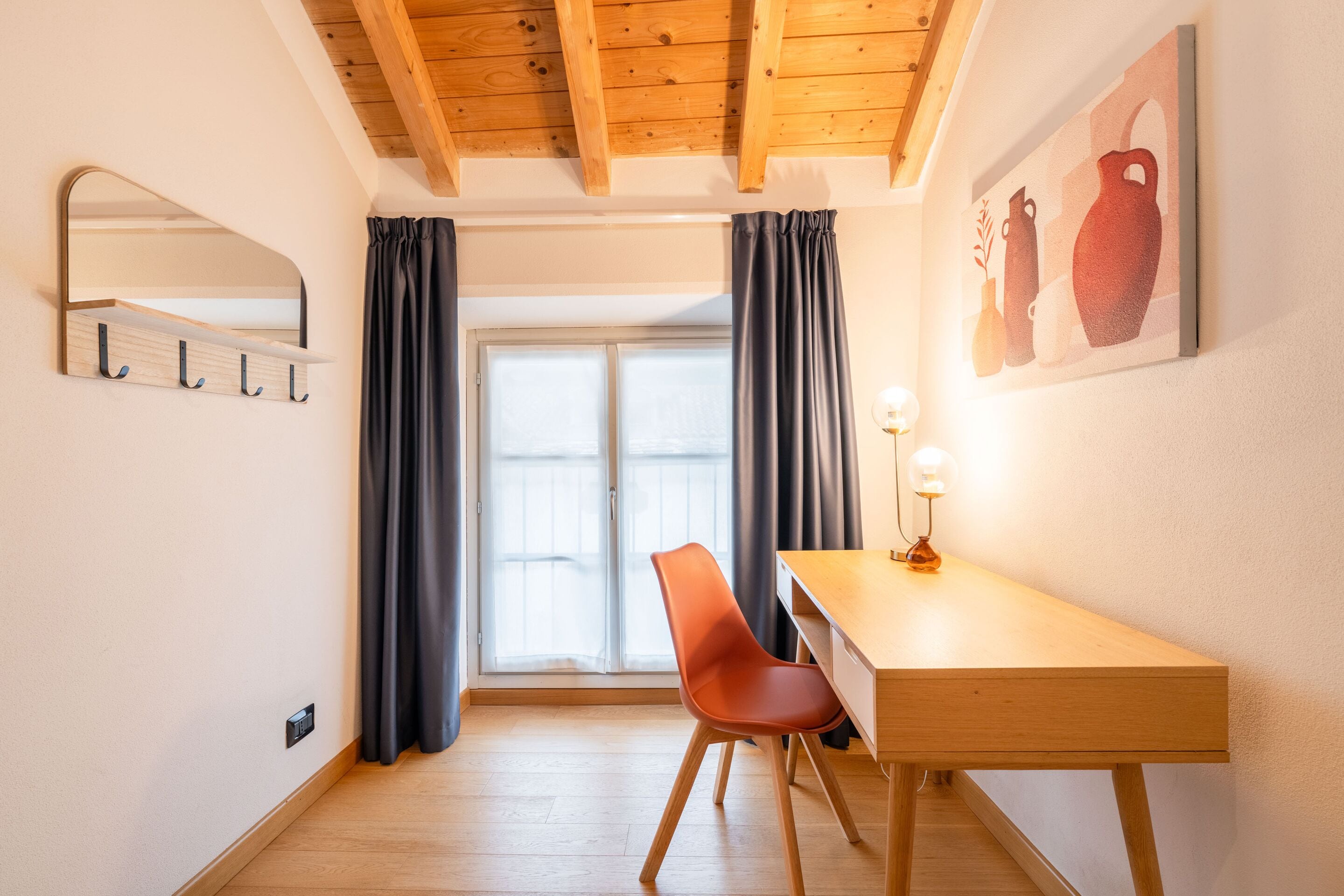 A room with a wooden desk, a chair, a mirror, and a painting on the wall.