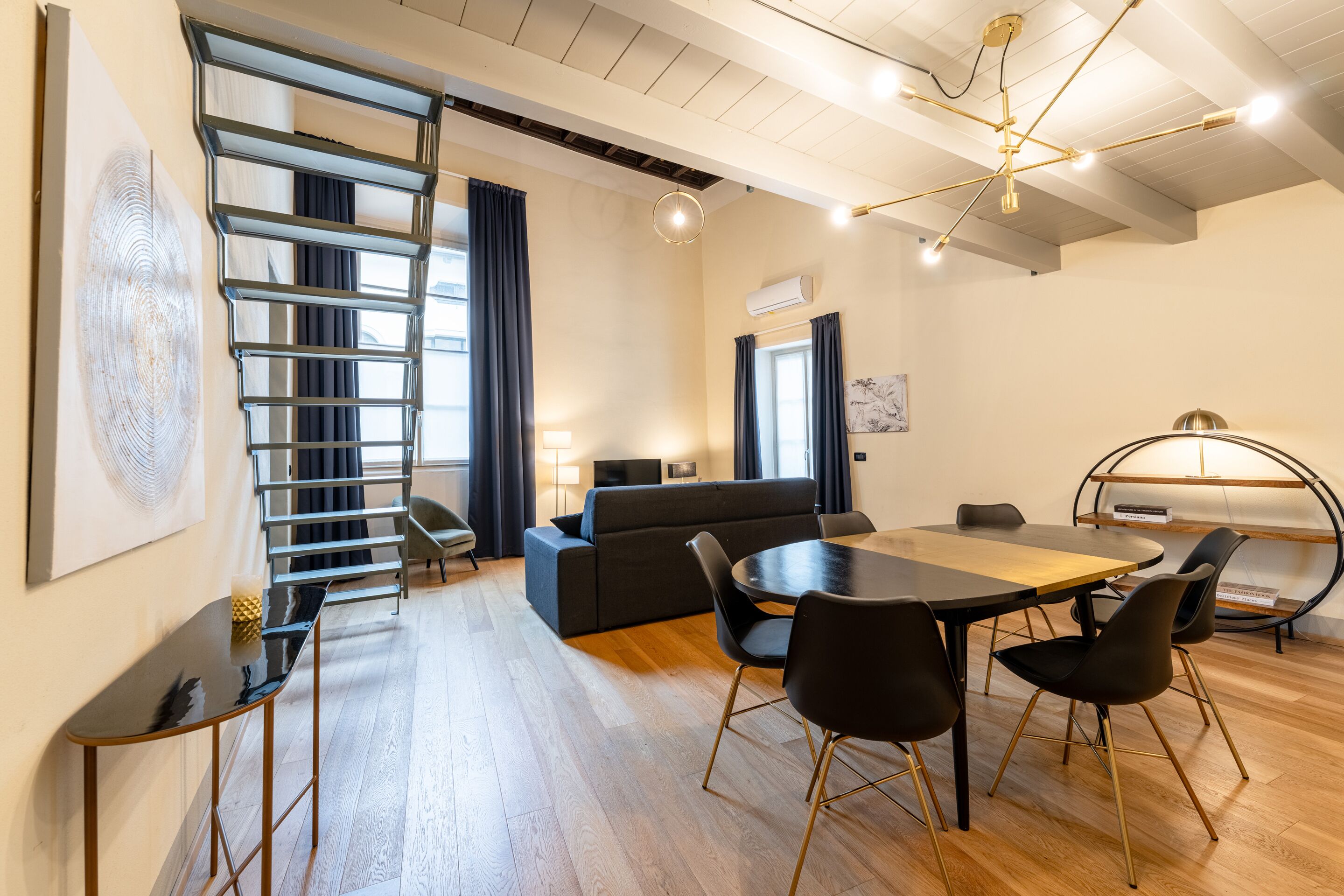 Ein modernes Wohnzimmer mit Holzboden, einem schwarzen Sofa, einem Esstisch mit Stühlen, einer Treppe und einer Deckenleuchte.