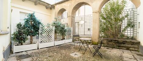 A courtyard with stone flooring, a small table with chairs, and a white fence with potted plants.