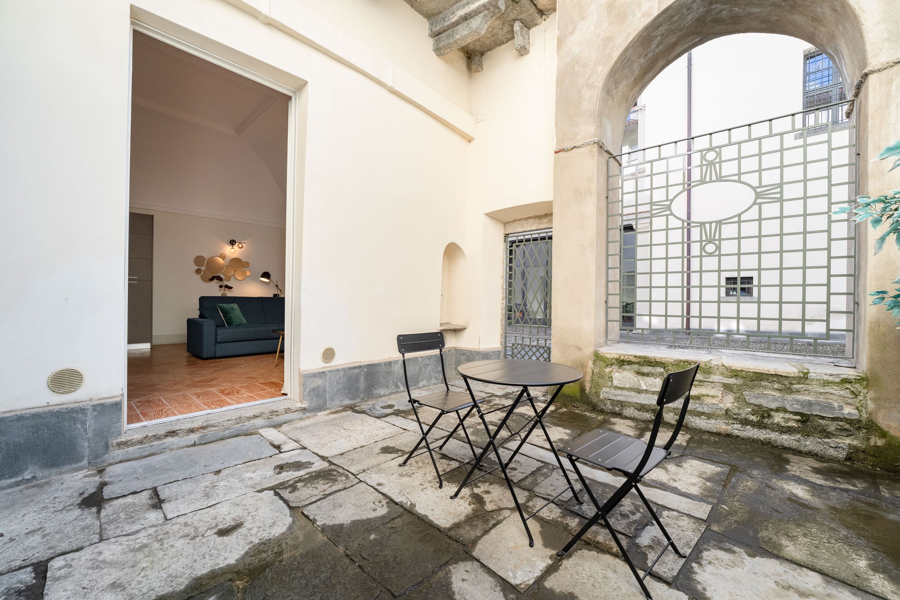 A courtyard with stone flooring, a dining table with chairs, and a view through a gate.