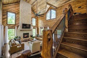 A rustic cabin interior with a stone fireplace, wooden staircase, and large windows. - Cabin | Hot Tub · Game Loft · Fire Pit (Broken Bow)