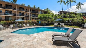 A pool area with lounge chairs and umbrellas, surrounded by palm trees and a multi-story building with balconies. - Kona Makai 3-204 (Kailua-Kona)