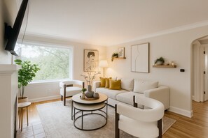 Een woonkamer met een witte bank, twee fauteuils, een salontafel en een aan de muur bevestigde televisie.