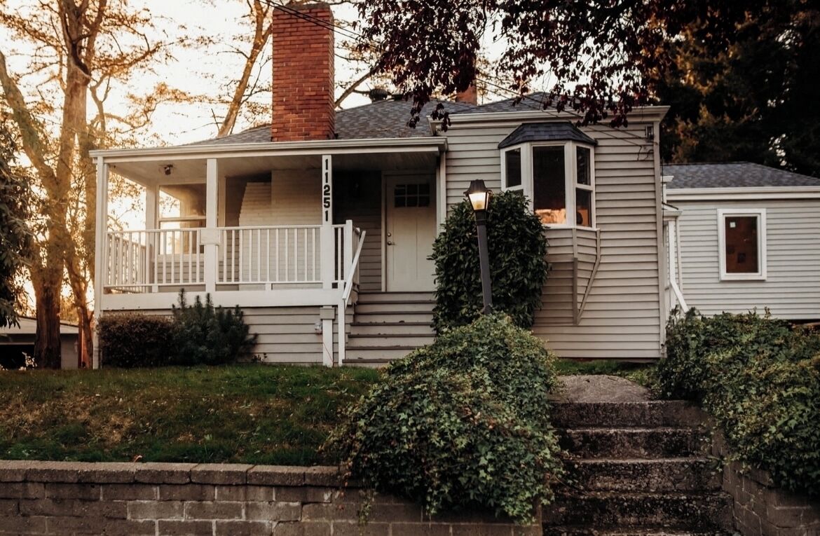 Une maison à deux étages avec un porche à l’avant, un petit garage attenant et une cheminée.