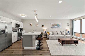 A modern kitchen with a central island, bar stools, a dining table, and a living area with a sofa and a coffee table.