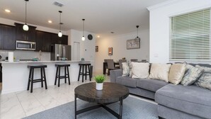 A modern living room with a grey sofa, a round coffee table, and a kitchen area with dark cabinets and stainless steel appliances. - 4br/3ba Private Pool Storey Lake Resort Best Location Near Disney! (Kissimmee)