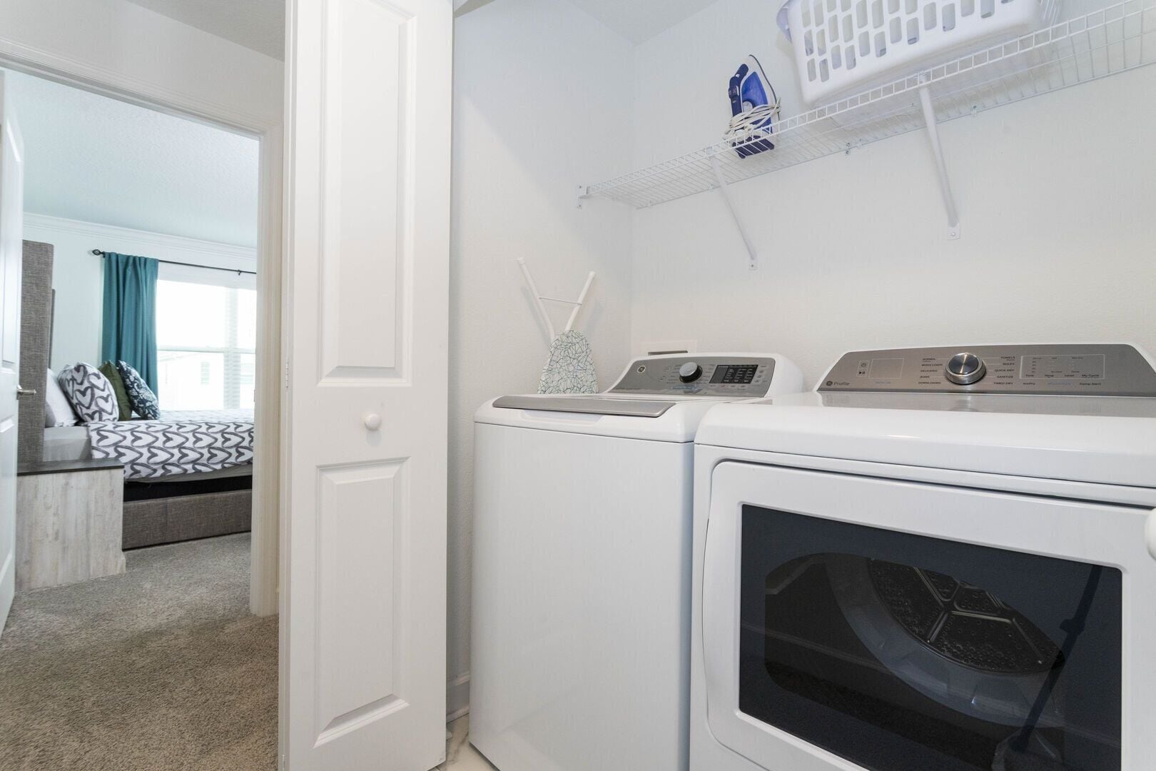 A laundry area with a washing machine and dryer, a shelf with laundry baskets, and a bed with patterned bedding.