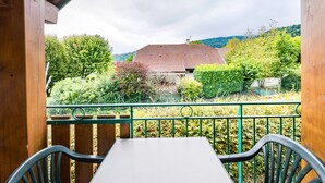 A wooden balcony with a table and chairs, overlooking a house with a red roof and a garden.