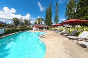 Ein Swimmingpool mit Liegestühlen und roten Sonnenschirmen, umgeben von Bäumen und blauem Himmel.