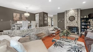 A modern living room with a brick fireplace, a dining area with a white table and chairs, and a bookshelf filled with books. - St Louis Co- Tranquil 2-bed/1 bath house -STL County-Sleeps 1-4 (Ballwin)