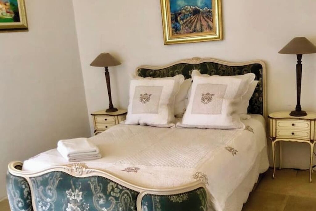 A classic four-poster bed with white linens and decorative pillows, flanked by two bedside tables with lamps, in a room with a framed painting on the wall.