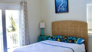 Un dormitorio con cama, ventilador de techo, ventana con cortinas, un cuadro en la pared y una mesita de noche con lámpara.